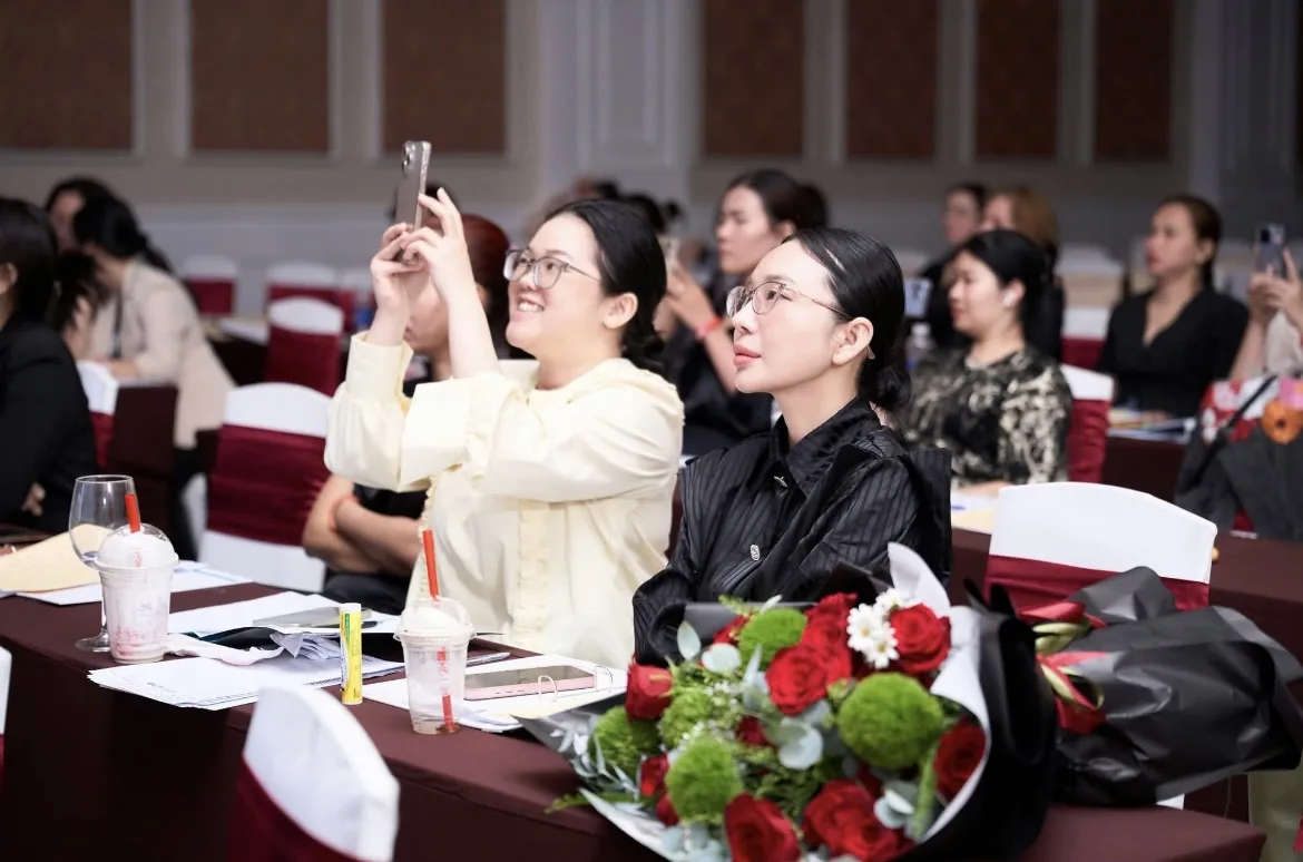 Học viên nữ người Việt đang giơ điện thoại chụp ảnh trong lớp học, ánh sáng cửa sổ tự nhiên, minh hoạ cách chụp ảnh cho spa không cần studio